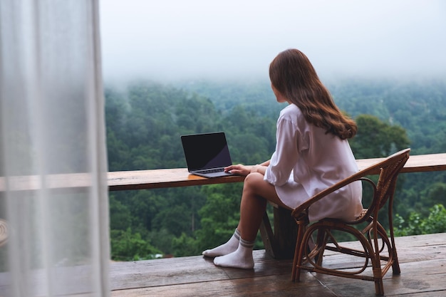 Achteraanzicht van een vrouw die een laptop gebruikt en eraan werkt terwijl ze op een balkon zit met een prachtig uitzicht op de natuur op een mistige dag