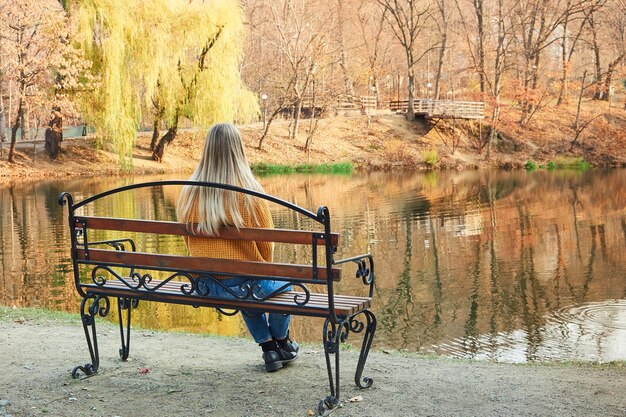 Achteraanzicht van een vrouw in een gele trui zittend op een bankje aan het meer in de herfst