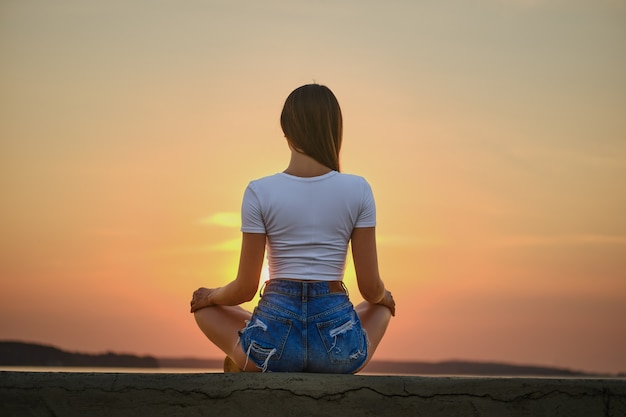 Achteraanzicht van een vrouw in meditatie pose op de pier tijdens de zomerzonsondergang