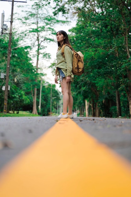 Achteraanzicht van een vrouwelijke wandelaar die over de asfaltweg in het bos loopt Toeristische vrouw met een rugzak die midden op de snelweg staat