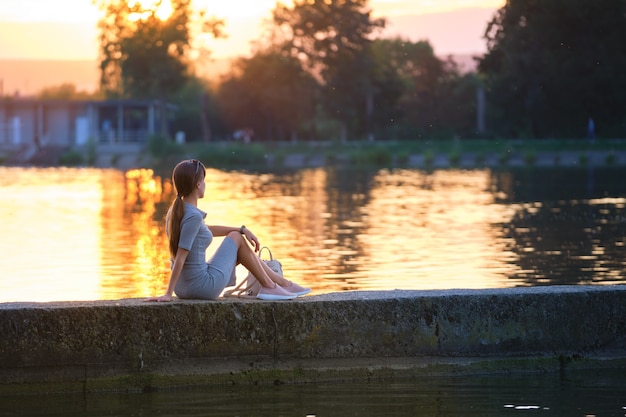 Achteraanzicht van eenzame vrouw zittend op de oever van het meer op warme avond. eenzaamheid en ontspanning concept.