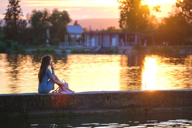 Achteraanzicht van eenzame vrouw zittend op de oever van het meer op warme avond. eenzaamheid en ontspanning concept.