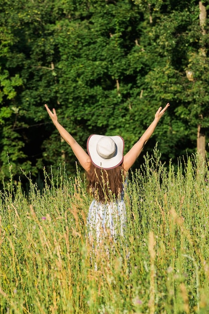 Achteraanzicht van gelukkige jonge vrouw met lang haar in hoed en jurk trekt haar armen omhoog tijdens een wandeling door het zomerbos op een zonnige dag Vreugde van het leven concept