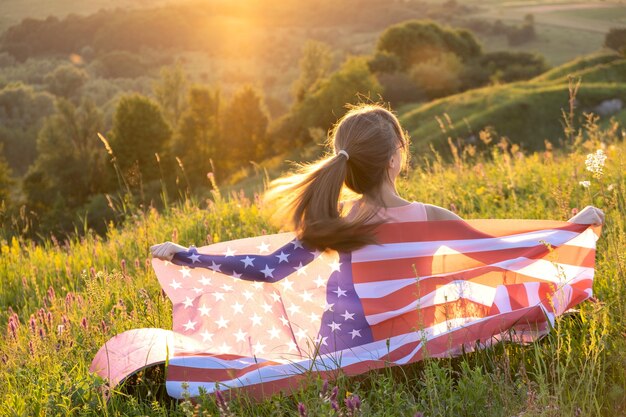 Achteraanzicht van gelukkige jonge vrouw poseren met usa nationale vlag buiten bij zonsondergang.