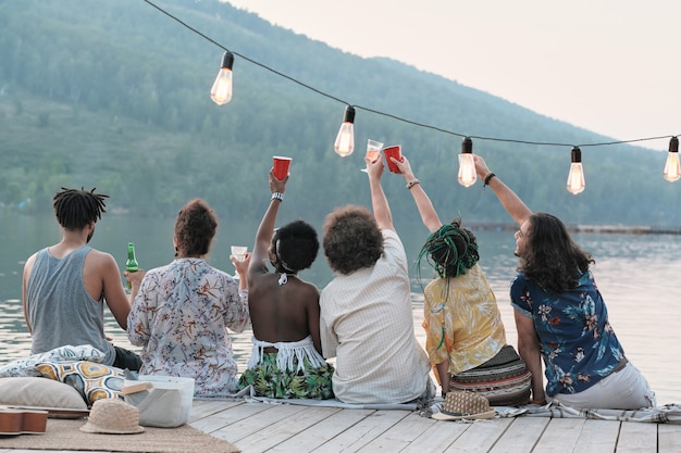 Achteraanzicht van groep vrienden roosteren met glazen bier en vieren hun zomervakantie zittend op een pier