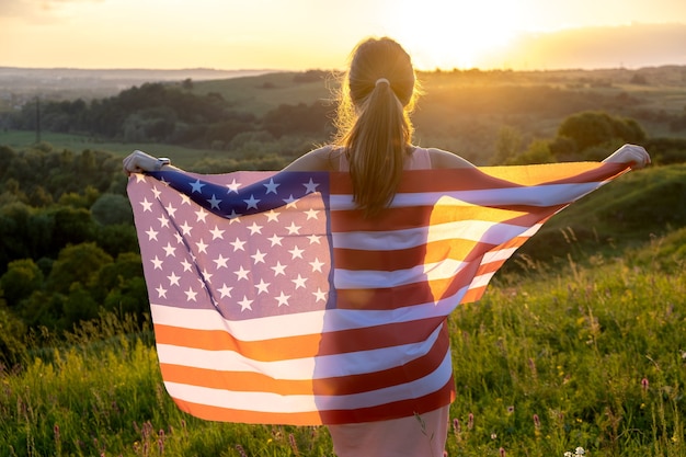 Achteraanzicht van het gelukkige jonge vrouw stellen met de nationale vlag van de VS die zich buiten bij zonsondergang bevindt