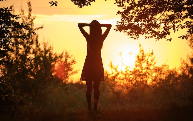 Achteraanzicht van het silhouet van een jonge vrouw die alleen door donkere bossen loopt met uitgestrekte handen in de zomeravond Het bereiken van je doel in het levensconcept