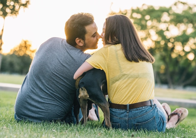 Achteraanzicht van jonge gelukkige paar zoenen met hun hond in het park. jong koppel verliefd zoenen met hun hond op gras.