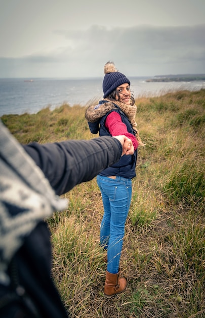 Achteraanzicht van jonge mooie vrouw met hoed en sjaal die de hand van de man vasthoudt en door een weide leidt. Volg mij naar het concept.