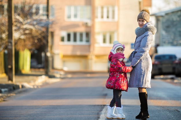 Achteraanzicht van jonge slanke aantrekkelijke vrouwenmoeder en de kleine dochter van het kindmeisje die in warme kleding holdingshanden op zonnige de winterdag lopen op vage stedelijke achtergrond.