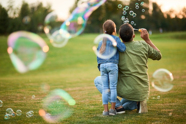 Achteraanzicht van jonge vader en zijn schattige kleine meisje die plezier hebben terwijl ze zeepbellen blazen in de