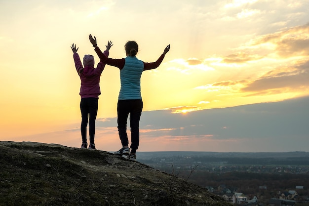 Achteraanzicht van jonge vrouw en kind meisje staande op met gras begroeide bergheuvel met opgeheven armen bij zonsondergang genieten van uitzicht op natuur familie toerisme tradities concept