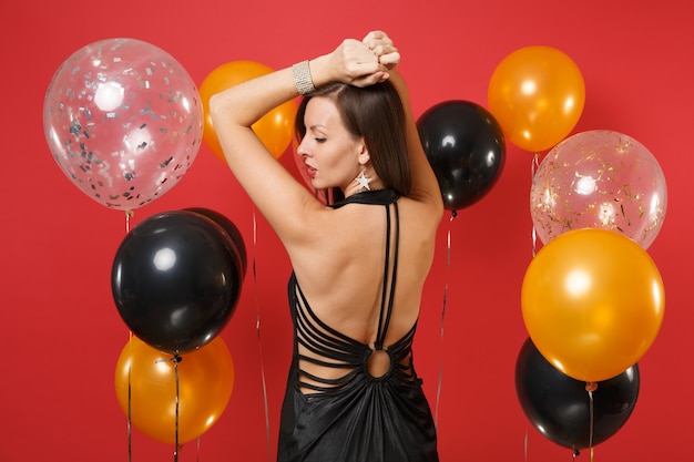 Achteraanzicht van jonge vrouw in zwarte jurk vieren, stijgende handen, opzij kijkend op heldere rode achtergrond luchtballonnen. St. Valentijnsdag, Gelukkig Nieuwjaar, verjaardag mockup vakantie feest concept.