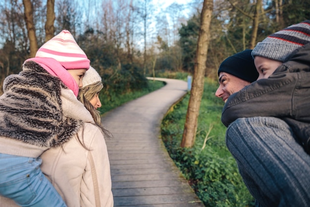 Achteraanzicht van lachende ouders die op de rug ritje geven aan gelukkige kinderen over een houten pad naar het bos