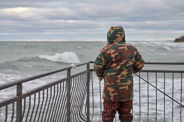 Achteraanzicht van mannelijke visser in camouflagepak die op de pier aan zee staat en een hengel vasthoudt. Mooie bewolkte hemel, horizon, zeegezicht.