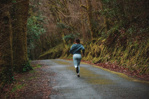 Achteraanzicht van onherkenbaar vrouwelijke atleet in sportkleding loopt langs asfaltweg in bos tijdens cardiotraining