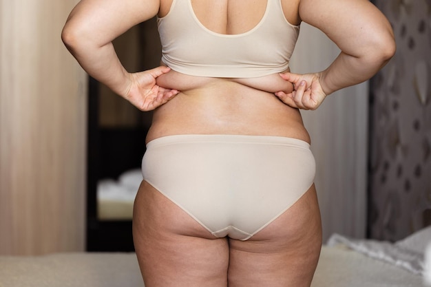Foto achteraanzicht van onherkenbare dikke mollige vrouw die in een beige beha-onderbroek in de buurt van bed staat en overmatig vet in de rug knijpt
