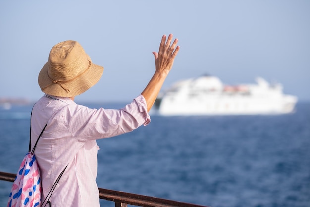 Achteraanzicht van senior vrouw met zomerhoed kijkend naar het vertrek van de veerboot zwaaiend met de hand