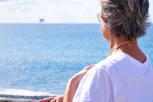 Achteraanzicht van senior vrouw wit gekleed leunend tegen een houten balkon met uitzicht op de zee