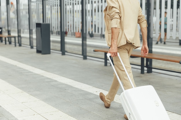 Achteraanzicht van succesvolle zakenman in beige pak lopen bij bushalte met witte koffer in de hand