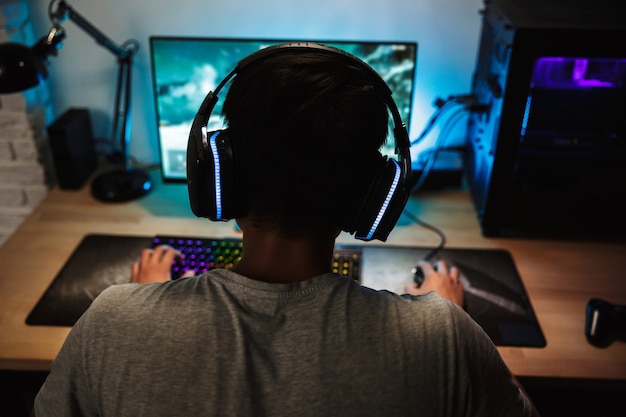 Achteraanzicht van tiener gamer jongen spelen van videospellen online op computer in de donkere kamer, hoofdtelefoon met microfoon dragen en kleurrijke toetsenbord met achtergrondverlichting gebruiken