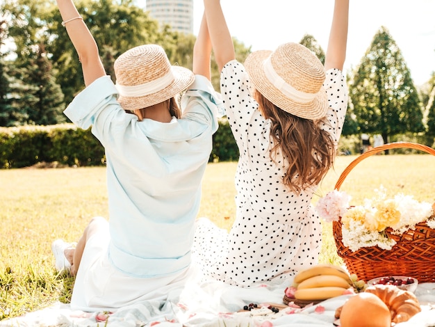 Achteraanzicht van twee jonge mooie glimlachende vrouwen in trendy zomerjurk en hoeden. Zorgeloze vrouwen die buiten picknicken.