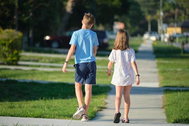 Achteraanzicht van twee jonge tienerkinderen, meisje en jongen, broer en zus die samen op landelijke straat lopen op een heldere zonnige dag Vakantietijdconcept