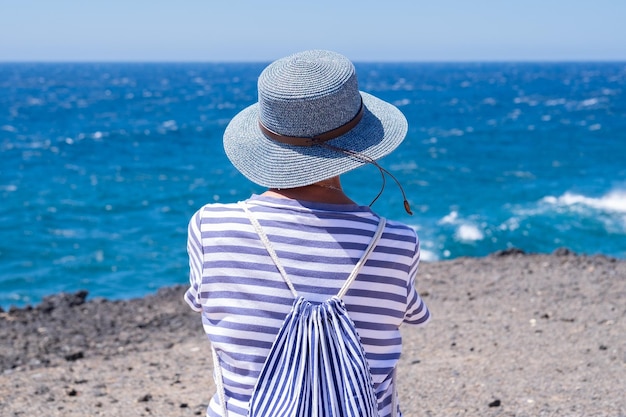 Achteraanzicht van volwassen vrouw gekleed in het blauw die de zee bewondert en naar de horizon over water kijkt rijpe dame die van ontspanning en vakantie geniet