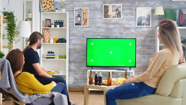 Achteraanzicht van vrienden die bier drinken en naar sport kijken op tv met groen scherm in de woonkamer.