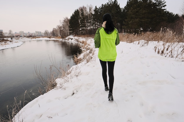 Achteraanzicht van vrouw in groene jas loopt langs de kust van de winter met lang droog gras