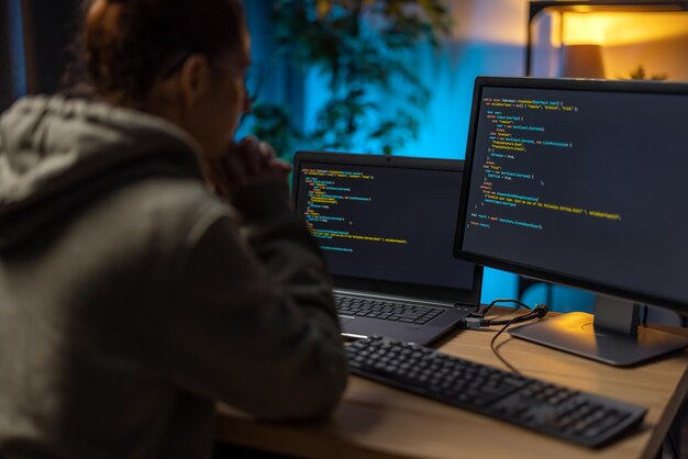 Achteraanzicht van vrouwelijke IT-specialisten die 's avonds thuis op twee moderne computers werken Blanke vrouw zit aan bureau met datacode op monitoren