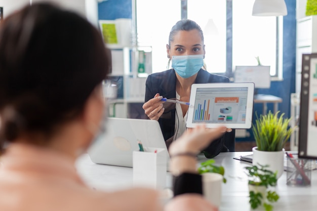 Achteraanzicht van zakelijke vrouwen met medisch gezichtsmasker die samenwerken bij managementpresentatie met behulp van tabletcomputer terwijl ze in het bedrijfskantoor zitten. Team met respect voor sociale afstand.