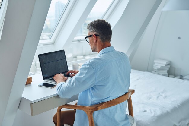 Achteraanzicht van zelfverzekerde volwassen man die op laptop werkt terwijl hij aan het bureau zit