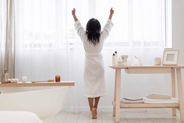 Achteraanzicht van zwarte vrouw in badjas die bij het raam in de badkamer staat