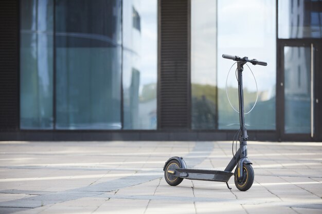 Achtergrondafbeelding van zwarte elektrische scooter staande op betegelde vloer tegen glazen gebouw in stedelijke omgeving, kopieer ruimte