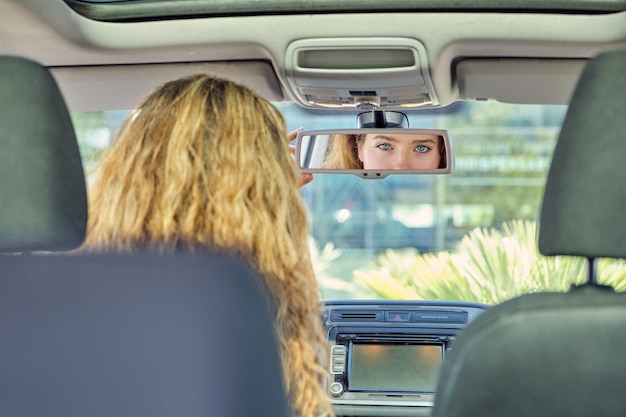 Foto achtergrondbeeld jonge vrouwelijke chauffeur met blauwe ogen en golvend haar die via de achteruitkijkspiegel naar de camera kijkt terwijl ze op de voorste stoel van de auto zit