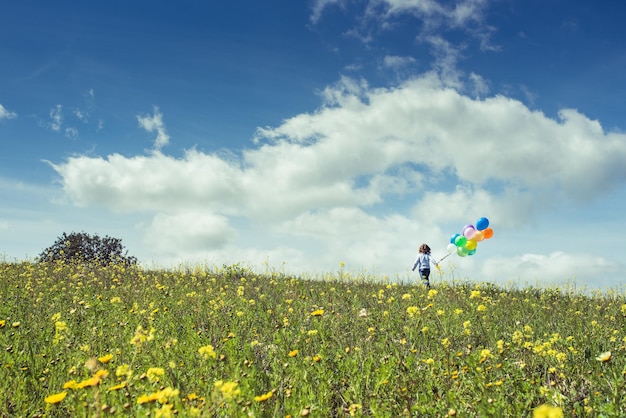 Achtergrondbeeld van anoniem kind met kleurrijke ballonnen terwijl hij op een zonnige dag op een bloeiende weide loopt onder een bewolkte hemel