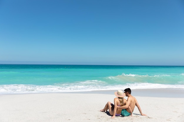 Achtergrondbeeld van een blank echtpaar dat op het strand ligt met blauwe hemel en zee op de achtergrond, omhelzend en wegkijkend