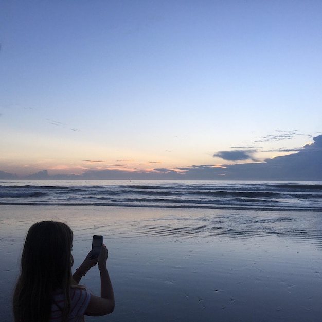 Foto achtergrondbeeld van een jonge vrouw die de zee tijdens de zonsondergang fotografeert met een mobiele telefoon