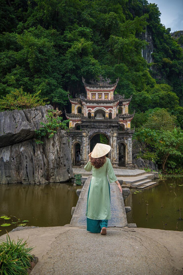 Achtergrondbeeld van een meisje dat Ao Dai draagt voor een parklandschap met een meer en een stenen brug. De ingang van de oude Bich Dong pagode complex Ninh Binh Vietnam reisbestemming
