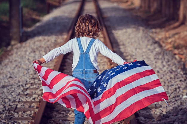 Achtergrondbeeld van een onherkenbaar kind in denim die op een zonnige dag op de spoorweg loopt met een zwaaiende Amerikaanse vlag