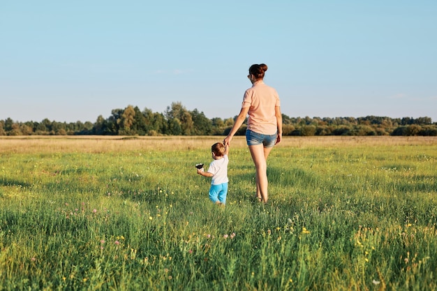 Achtergrondbeeld van jonge moeder en haar jonge dochter die op het gras lopen in de zomergroene weide met groene bomen op de achtergrond en samen genieten van hete zonnige dagen