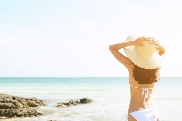 Achterkant jonge vrouw huidskleur in zonnehoed en bikini staand met haar armen naar haar hoofd geheven genieten op het strand vakantie reizen en genieten van het leven op zee op zoek naar uitzicht op strand oceaan op warme zomerdag