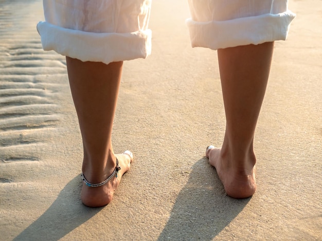 Achterkant van blootsvoets vrouw in witte broek met gevouwen benen staande op schoon zandstrand in het warme zonlicht bij zonsondergang 's avonds zomer reizen vrijetijdsbestemming en vakantie concepten