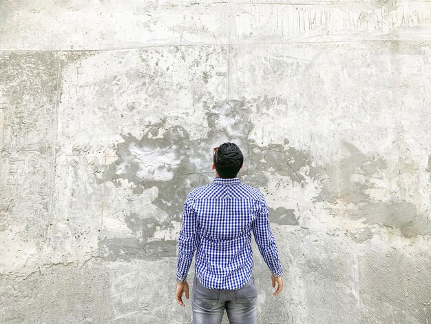 Achterkant van jonge man in geruit blauw shirt en zonnebril staande tegen betonnen grijze muur. kijken naar achtergrond lege copyspace.