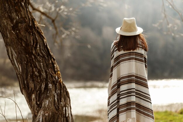 Achterkant vrouw hipster hoed en poncho dragen bij boom in de buurt van rivier amerikaanse outfit boho concept