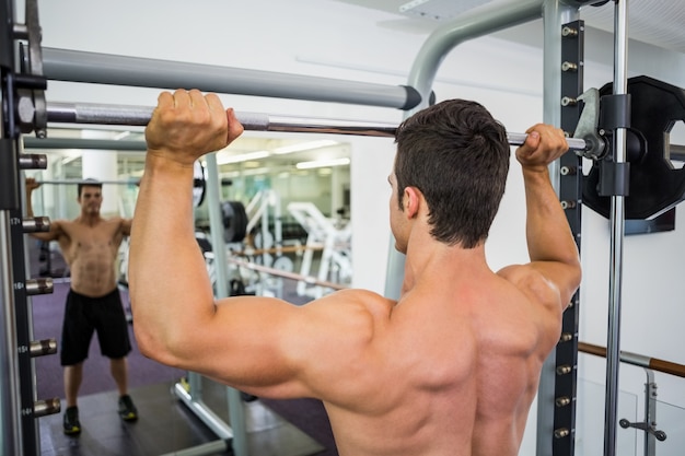 Achtermening van een shirtless spiermens die barbell in gymnastiek opheffen