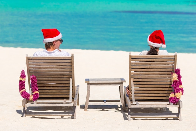 Achtermening van jong paar die in Kerstmanhoeden op tropisch strand tijdens Kerstmisvakantie ontspannen