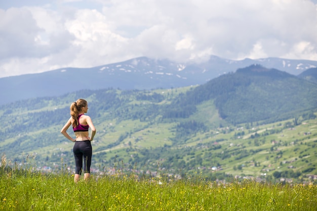 Achtermening van slanke jonge vrouw die zich op grasrijke vallei op achtergrond van groene bergen op zonnige de zomerdag bevinden.