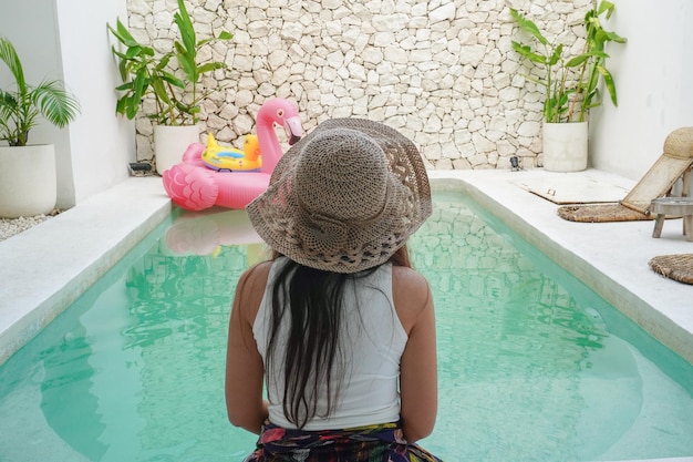 Achterportret van een jonge Aziatische vrouw met een witte top met een lichte crème gekleurde hoed die bij het zwembad in een villa zit te ontspannen en te genieten van het uitzicht op de zomervakantie